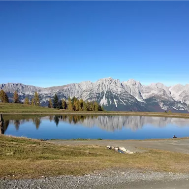 A tranquil lake reflects the surrounding mountains and the clear blue sky. In the foreground, green meadows and some trees are visible.