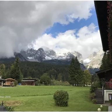 A picturesque landscape with majestic mountains and a cloudy sky. In the foreground, there is a green meadow and traditional wooden chalets.