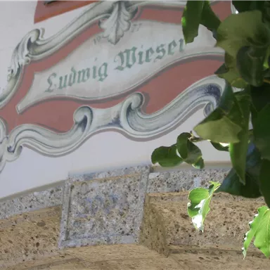 A decorative sign with the name "Ludwig Wieser" is mounted on a wall. Green plants frame the picture.