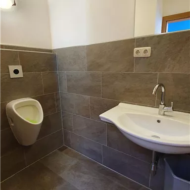 A modern bathroom with gray tiles. It features a urinal, a sink, and bright lighting.