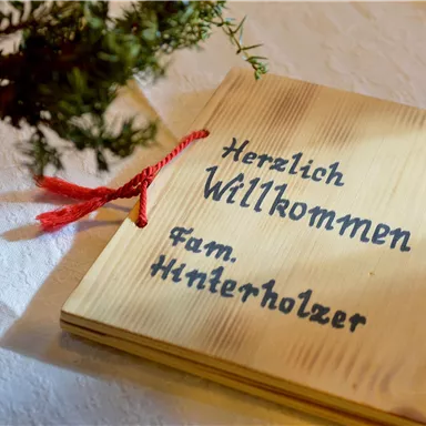 Ein Willkommensschild aus Holz mit der Aufschrift „Herzlich Willkommen Fam. Hinterholzer“. Daneben steht ein kleiner Topf mit einer Pflanze.