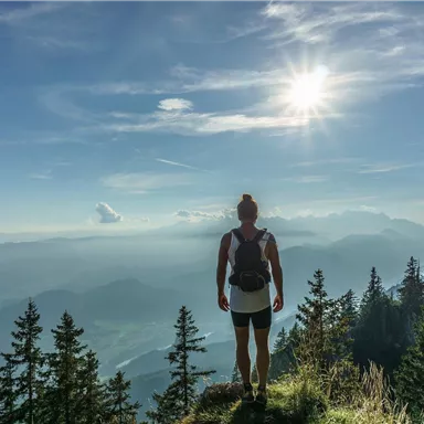 A person is standing on a mountain and looking into the distance. The sun is shining clearly over a picturesque landscape with trees and hills.