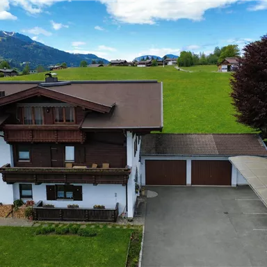 A charming alpine house with a large garden and plenty of green space. In the background, gentle hills and a blue sky can be seen.