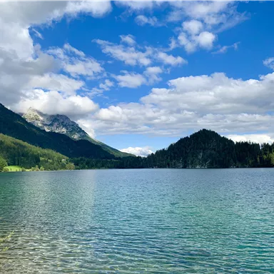 Ein klarer See umgeben von Bergen und Bäumen. Der Himmel ist blau mit einigen Wolken.