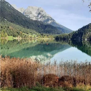 Ein ruhiger See umgeben von Bergen und grünen Wäldern. Das Wasser spiegelt die Landschaft wider und der Himmel ist leicht bewölkt.