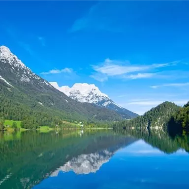 Ein klarer See, umgeben von majestätischen Bergen und üppigem Grün. Der Himmel ist strahlend blau und spiegelt sich im Wasser.