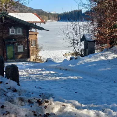 A winter landscape with a frozen lake and snow. In the foreground, there is a traditional wooden house.
