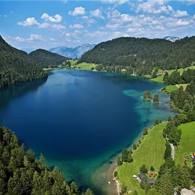 A clear lake surrounded by lush forests and gentle hills. The sky is blue with a few white clouds.