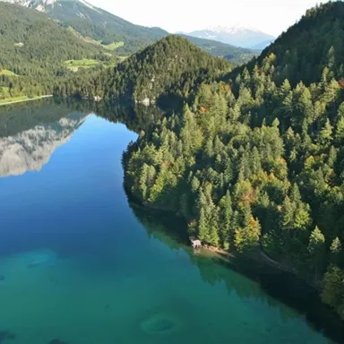 Ein klarer See umgeben von hohen Bäumen und Bergen. Die Landschaft spiegelt sich im ruhigen Wasser.
