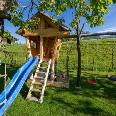 Ein schöner Spielplatz mit einem Baumhaus und einer Rutsche. Schaukeln und eine grüne Wiese umgeben den Bereich.