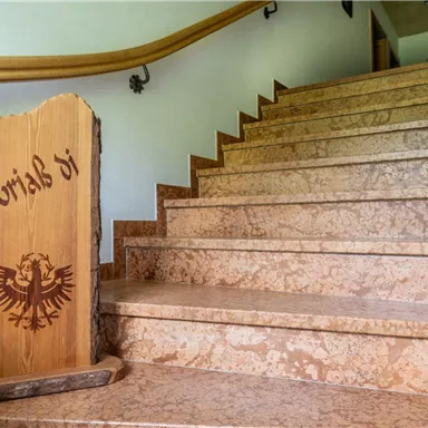 Eine Treppe aus hellem Stein mit einem Holzschild, das den Text "Grüß di" zeigt. Die Wände sind in sanften Farben gehalten und ein Geländer aus Holz verläuft entlang der Treppe.