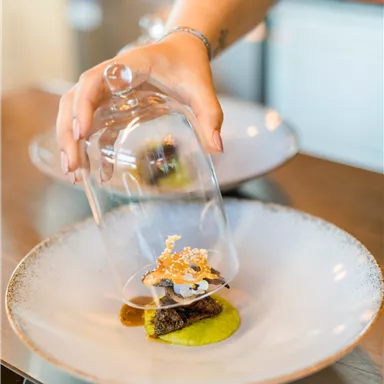 A high-quality dish is presented on a plate. A hand lifts a glass dome to reveal the food.