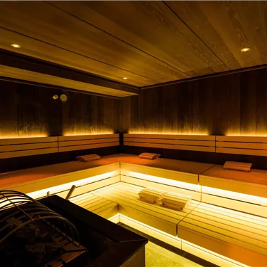 A modern sauna interior with warm, soft lighting. The wood paneling and benches create a relaxing atmosphere.