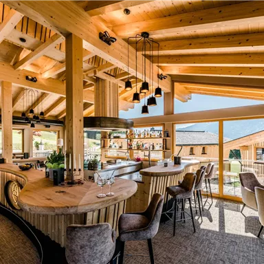 A modern restaurant with wooden construction and large windows. The space is bright and inviting, with a central bar and stylish seating.
