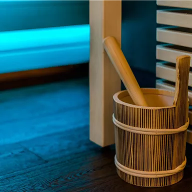 A wooden bucket with a wooden spoon is next to a sauna. In the background, gentle blue light elements can be seen.