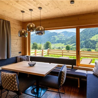 A cozy dining area in a wooden house with a view of the green mountains. Large windows let in a lot of light and offer a beautiful view of the landscape.
