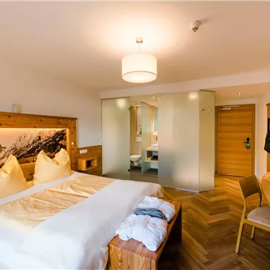 A modern hotel room with wooden decor and large windows. The bed is cozy and the bathroom is separated by a glass wall.