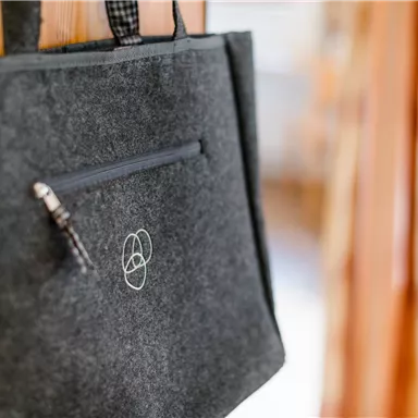 A gray felt bag hangs on a wall. It has a zipper and a simple logo.