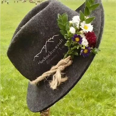 A gray hat with a floral decoration stands on a stick in a meadow. In the background, green fields and mountains are visible.