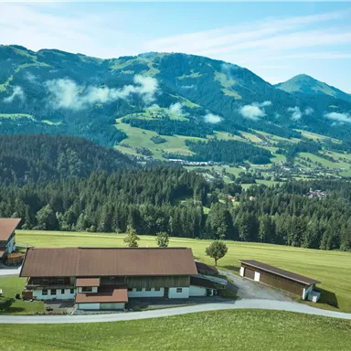 Eine malerische Landschaft mit sanften Hügeln und bewaldeten Bergen. Im Vordergrund sind rustikale Gebäude auf einer grünen Wiese zu sehen.
