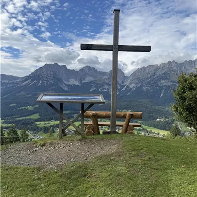 Eine Aussicht auf die Berge mit einem Holzkreuz im Vordergrund. Eine Tisch-Bank-Kombination lädt zum Verweilen ein.