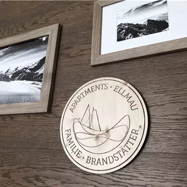 A wooden sign with the inscription "Apartments Ellmau Familie Brandstätter" hangs on a wooden wall. Next to it are two framed black-and-white pictures of landscapes.
