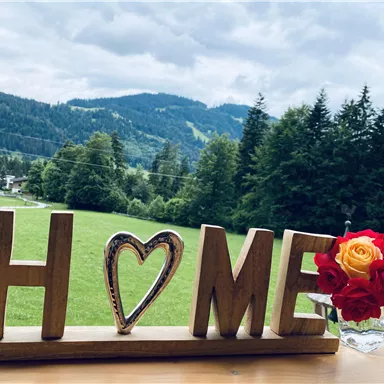 Eine Holzdekoration mit dem Wort "HOME" steht im Vordergrund. Im Hintergrund sieht man eine grüne Landschaft mit Bergen und Bäumen.