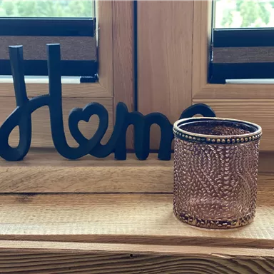 Ein dekoratives „Home“-Schild aus Holz steht neben einem eleganten Glasbehälter auf einem Holzfensterbrett. Die Szene strahlt Gemütlichkeit und Zuhause-Feeling aus.