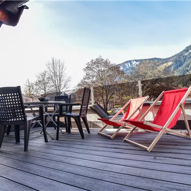 A terrace with a table and four chairs, including two red lounge chairs. In the background are trees and mountains under a clear sky.