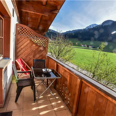 A cozy balcony with a table and chairs offers a view of green meadows and mountains in the background. The sun shines on the relaxing surroundings.