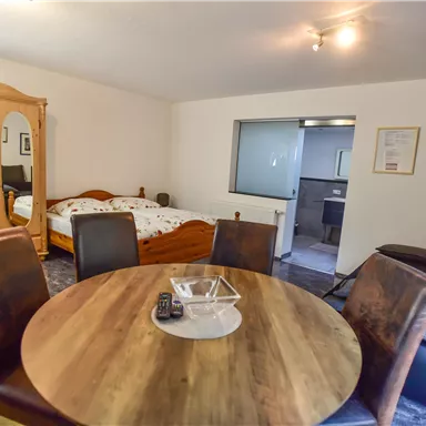 A cozy room with a round dining table and chairs. In the background, a bed and a mirrored wardrobe are visible.