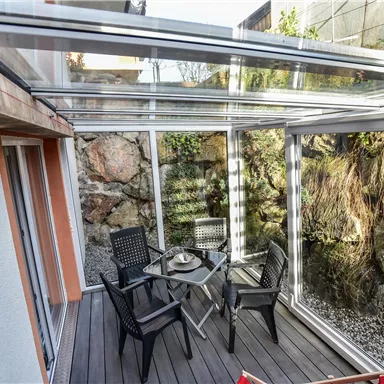 A modern conservatory with glass walls and a wooden floor. In the center stands a table and chairs, surrounded by a natural stone wall.