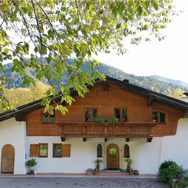 Ein traditionelles alpenländisches Haus mit Holzverkleidung und einem blühenden Balkon. Umgeben von grünen Bäumen und sanften Hügeln.