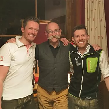 Three men pose in traditional clothing in front of a window. They are smiling and seem to be enjoying a sociable atmosphere.