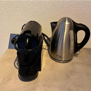 A black fully automatic coffee machine stands next to a silver kettle on a gray surface. Both devices are suitable for preparing beverages.