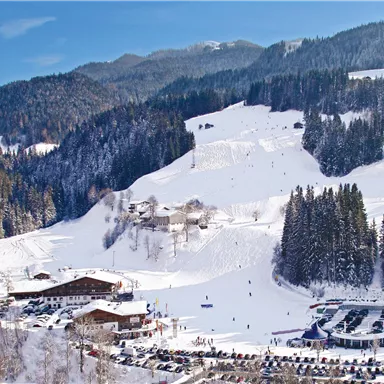 A picturesque winter landscape with snow-covered mountains and a ski resort. In the foreground, there is a parking area and some buildings.