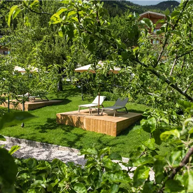 Ein ruhiger Garten mit Liegen auf Holzterrassen. Umgeben von grünen Bäumen und frischem Gras.