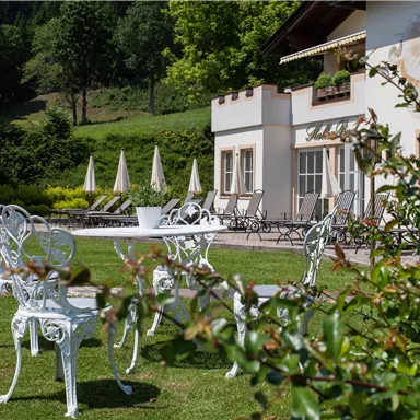 Ein schöner Garten mit weißen Stühlen und Sonnenschirmen. Im Hintergrund ist ein Gebäude mit Fenstern und grünen Bäumen.