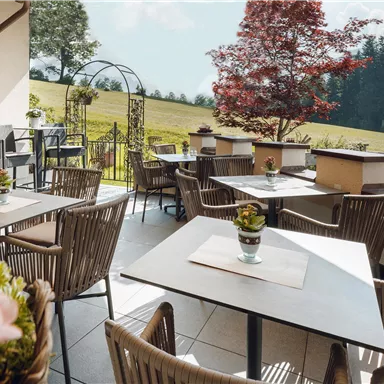 Eine gemütliche Terrasse mit modernen Tischen und Stühlen. Im Hintergrund ist eine grüne Wiese und Bäume zu sehen.
