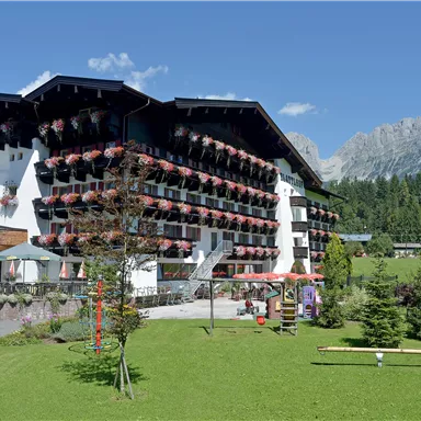 A picturesque hotel building with balconies adorned with flowers. In the background, green meadows and impressive mountains can be seen.