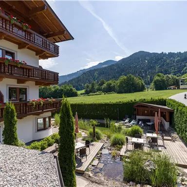 Ein charmantes Hotel mit einem Garten, umgeben von Bergen und grünen Wiesen. Die Balkone sind mit Blumenkästen geschmückt und der Himmel ist blau.