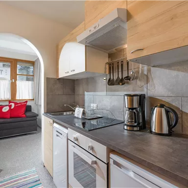 A modern kitchen with wood and gray stone surfaces. In the background, a cozy living room with a sofa and a colorful rug is visible.