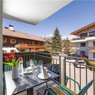 A beautiful balcony with a table on which flowers and glasses are placed. In the background, modern and traditional buildings as well as trees can be seen.