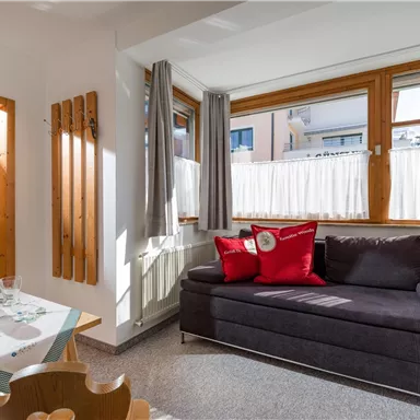 A bright living room with large windows and a cozy sofa. A dining area with a table and fresh flowers complements the space.