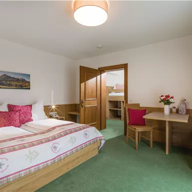 A cozy room with a large bed and wooden details. There is a dining area and a balcony with a view of the mountains.