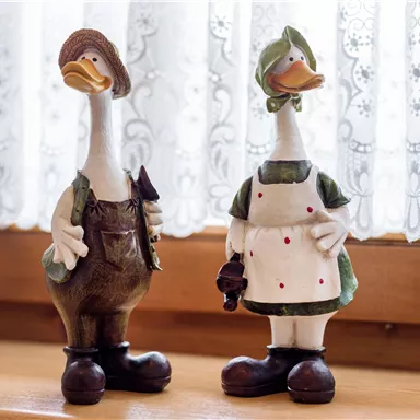 Two decorative duck figures are standing on a windowsill. One is wearing overalls and the other is wearing a dress with an apron.
