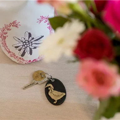 A bunch of keys with an interesting keychain is lying on a table. In the background, colorful flowers can be seen.