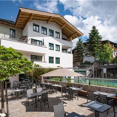 A modern building with an outdoor area and a pool is visible. The terrace is furnished with tables and chairs, surrounded by trees and nice weather.