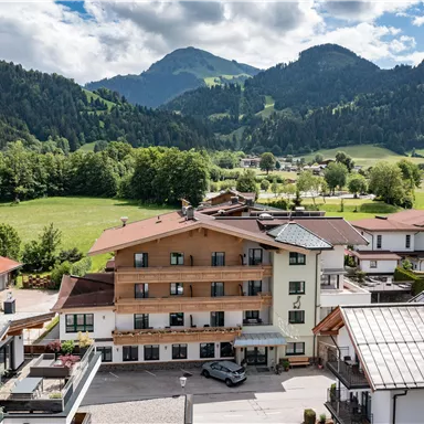 A picturesque mountain village with modern houses and many trees. In the background, impressive mountains rise under a partially cloudy sky.