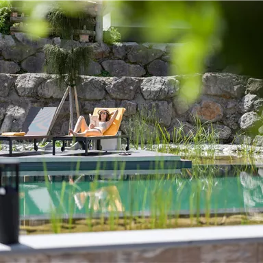 A relaxed person is lying on a lounger by the water. Surrounded by green plants and stones, the place exudes peace and relaxation.